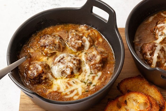 Two bowls of Taste of Home French Onion Meatballs in black bowls on a wooden cutting board, with toasted baguette slices