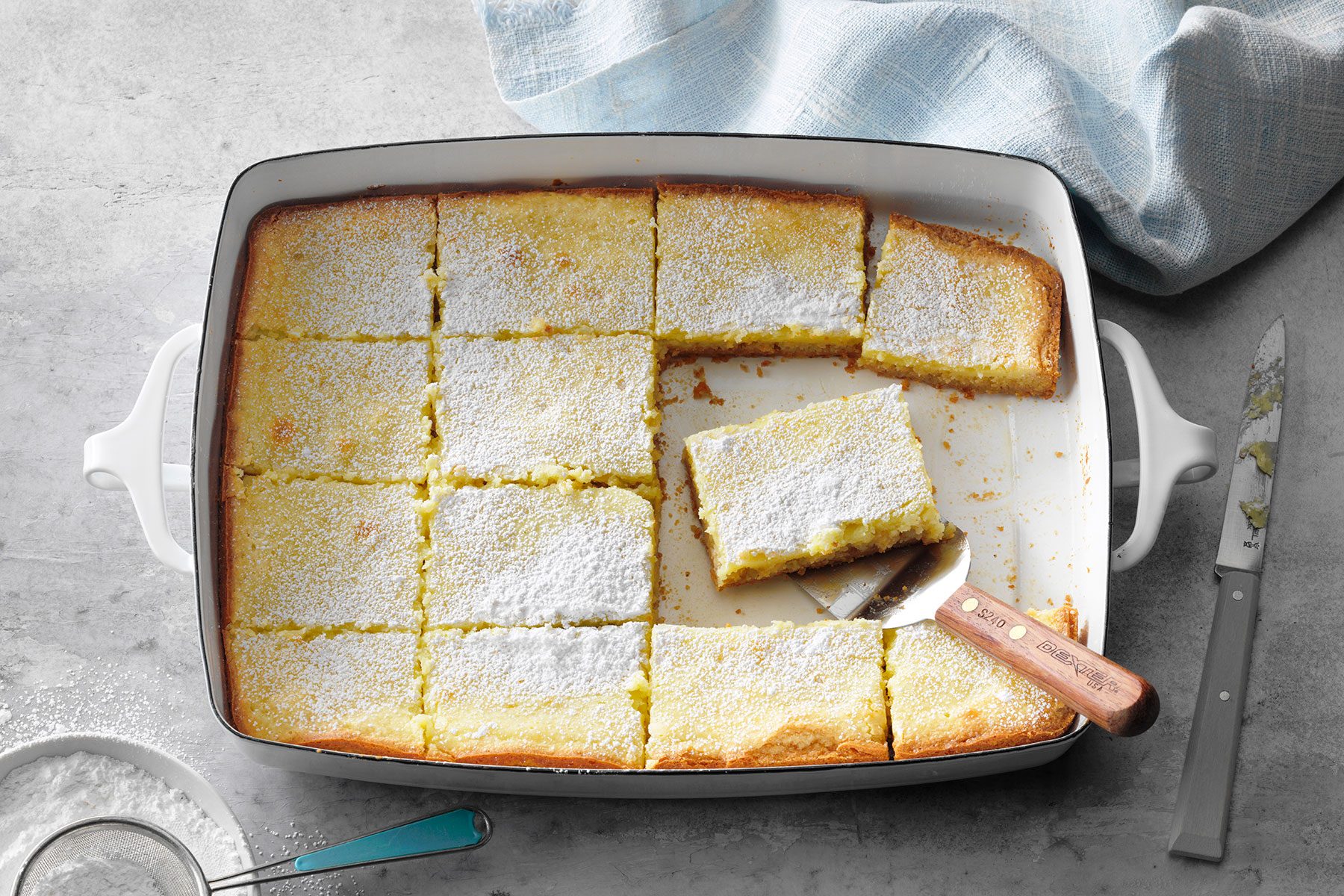A baking dish filled with square slices of cake, dusted with powdered sugar. One slice is partially lifted by a spatula. A knife and a blue cloth are nearby on the gray surface.