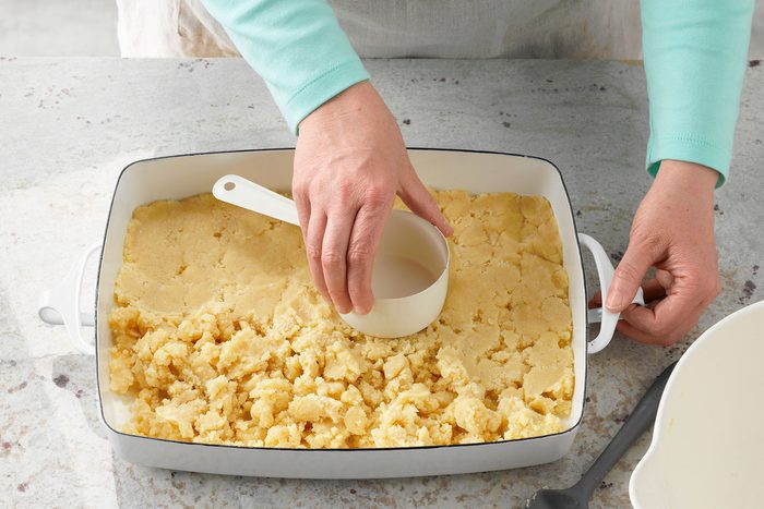 A person in a light blue sleeve presses a measuring cup onto a crumbly dough mixture inside a white rectangular baking dish on a speckled countertop.
