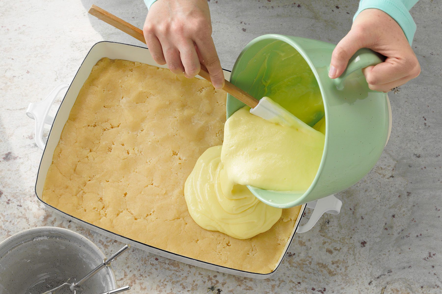 A person is pouring creamy yellow batter from a green mixing bowl into a rectangular baking dish containing a dough-like base. The image shows a hand using a spatula to scrape the batter out. The scene is set on a marble counter.