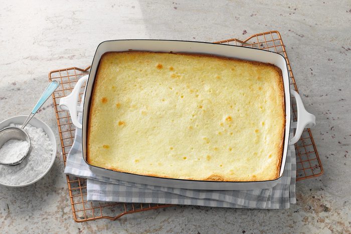 A golden-brown pudding in a white rectangular baking dish, placed on a checkered cloth and wire rack. A bowl of powdered sugar with a spoon is beside the dish on a light stone surface.