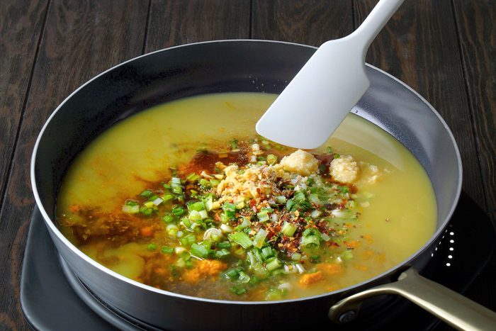 A pan on a stove filled with a yellow broth, topped with chopped green onions, garlic, chili flakes, and spices. A white spatula is positioned on the broth, suggesting stirring. The background is a dark wooden surface.