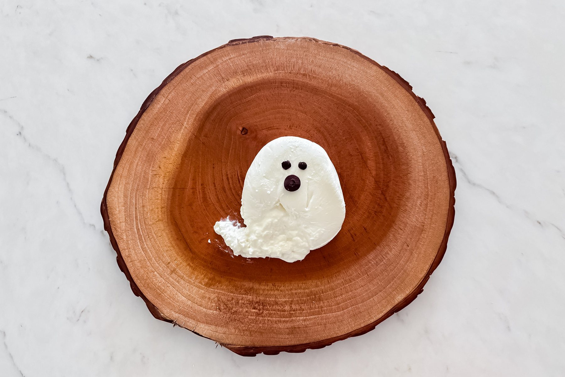 A dessert resembling a small ghost, made of a dollop of white cream with two black eyes and a black mouth, sits on a round wooden plate. The plate is placed on a white marble surface.