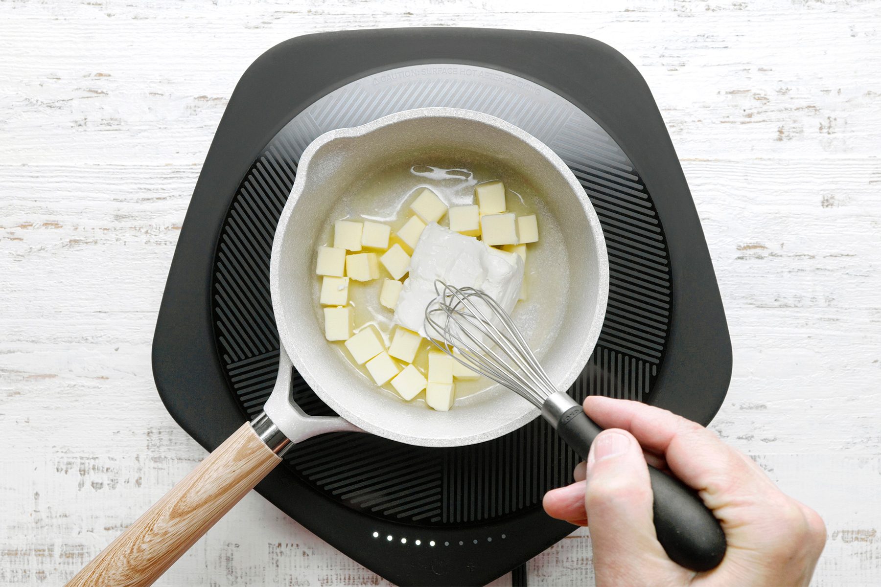 overhead shot of a white saucepan on an electric induction cooktop contains a mixture of melted butter and a liquid, likely water or milk, The butter is a pale yellow color and is bubbling slightly, A whisk is being used to stir the mixture together;