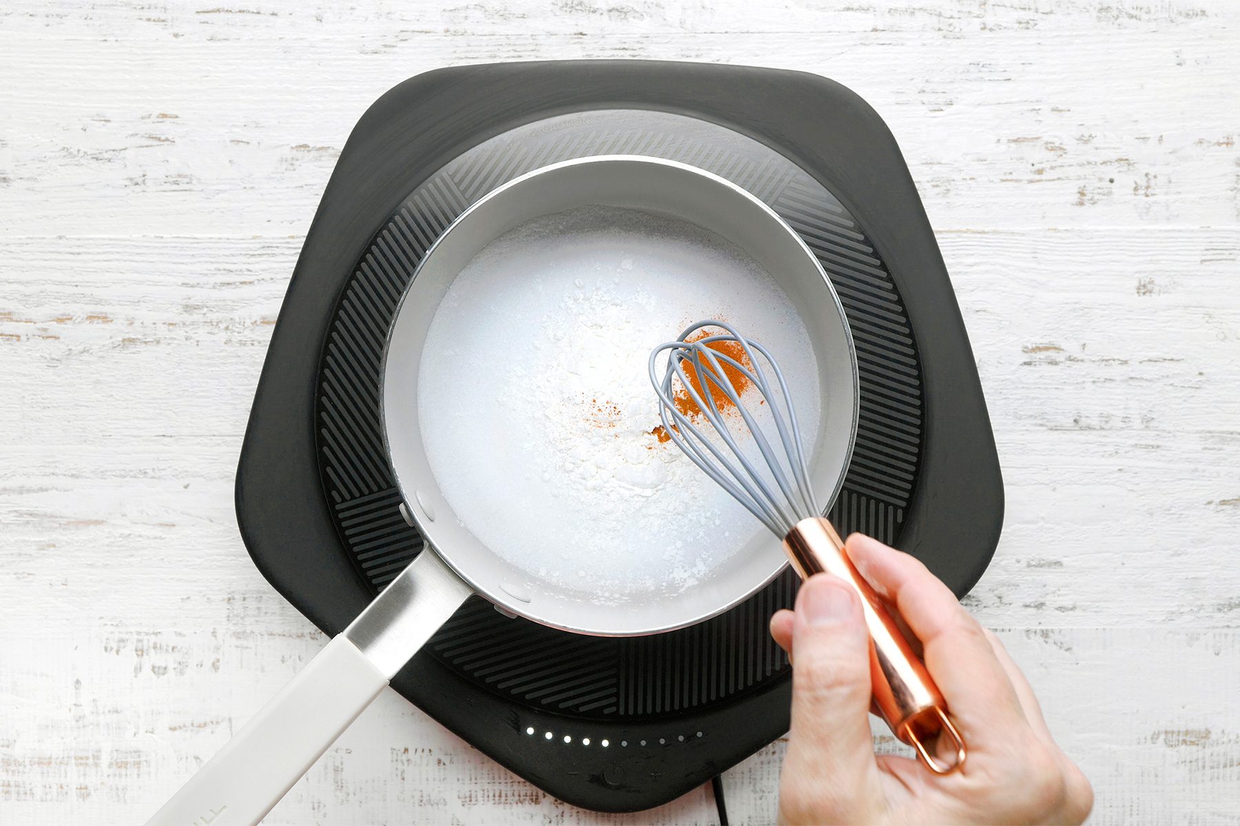 overhead shot of a saucepan on an electric induction cooktop contains a liquid mixture, A whisk is being used to stir the mixture, which has a sprinkle of cinnamon on top, The liquid appears to be a pale yellow color and is slightly bubbling;