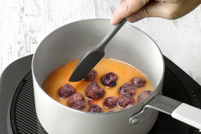 3/4th shot of a saucepan on an electric induction cooktop contains a mixture of liquid and whole cherries, The liquid is a reddish-orange color and is bubbling slightly, A spatula is being used to stir the mixture, ensuring the cherries are evenly coated in the liquid;