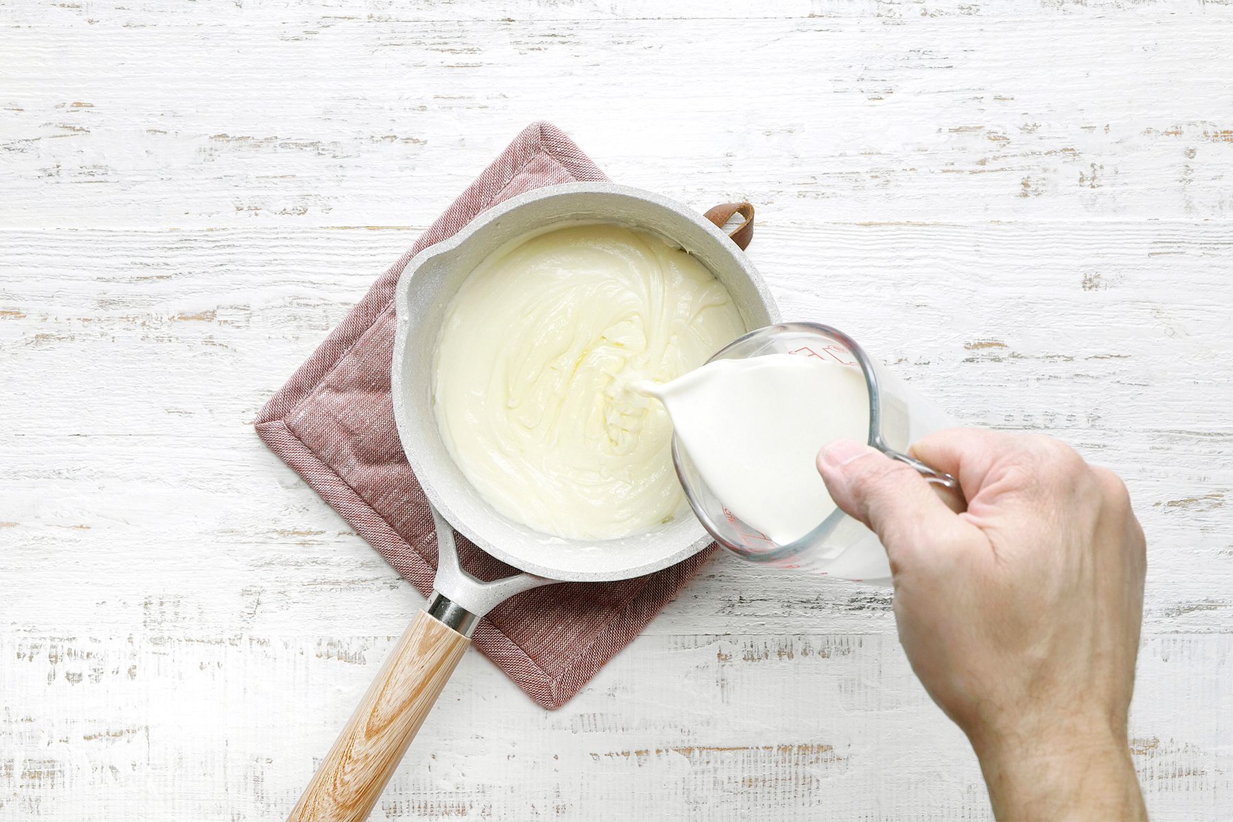 overhead shot of a white saucepan is filled with a creamy, pale yellow sauce, A person is pouring a liquid, likely milk or cream, into the saucepan, The liquid is creating ripples and swirls in the sauce;