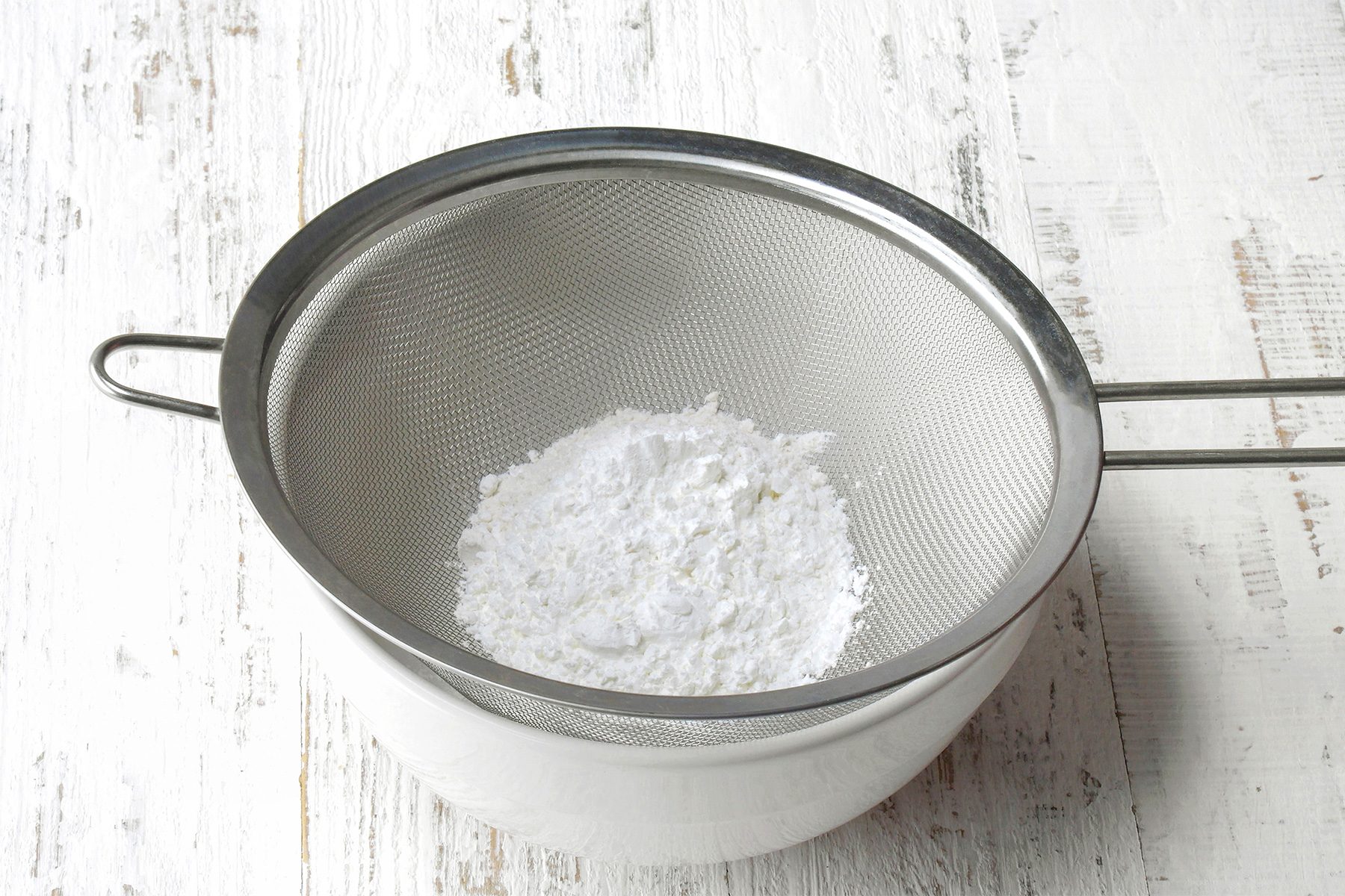 3/4th shot of a white bowl is placed underneath a metal mesh strainer, A fine white powder, likely powdered sugar, is resting in the strainer, The powder appears to be sifted through the mesh, as some particles have fallen through into the bowl below;