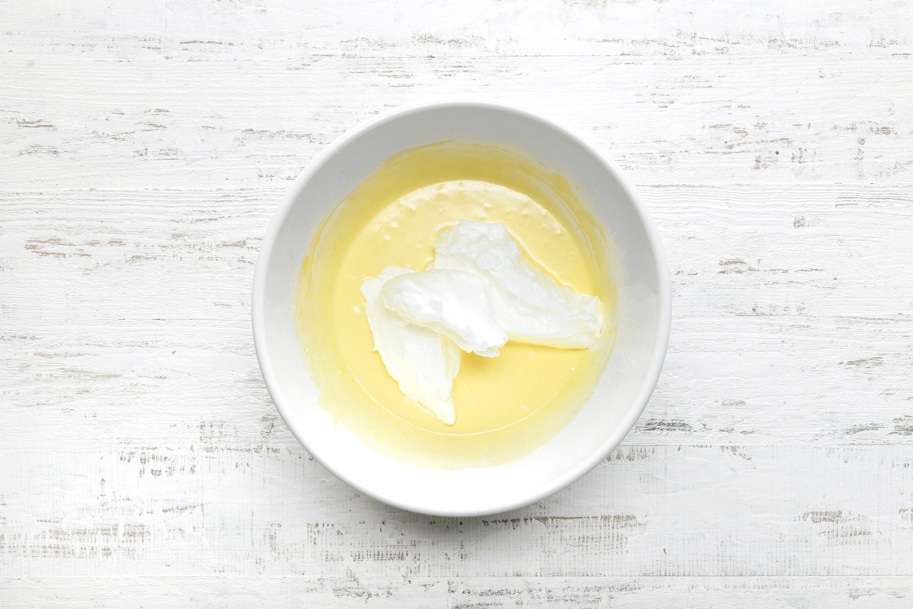 overhead shot of a white bowl containing a mixture of egg yolks and a light yellow liquid, Mounds of whipped egg whites are folded into the mixture, The bowl is placed on a wooden surface;