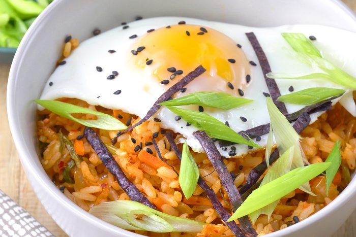 close shot of a bowl of Kimchi Fried Rice is showcased, featuring a vibrant mixture of rice, kimchi, vegetables, and a fried egg on top, The rice is tinged with a reddish hue from the kimchi;