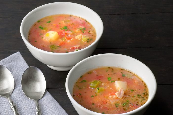 Two white bowls filled with vegetable soup, featuring chunks of potato, tomato, celery, and herbs, are placed on a dark wooden table. Two silver spoons rest nearby on a folded gray napkin. The soup appears fresh and colorful.