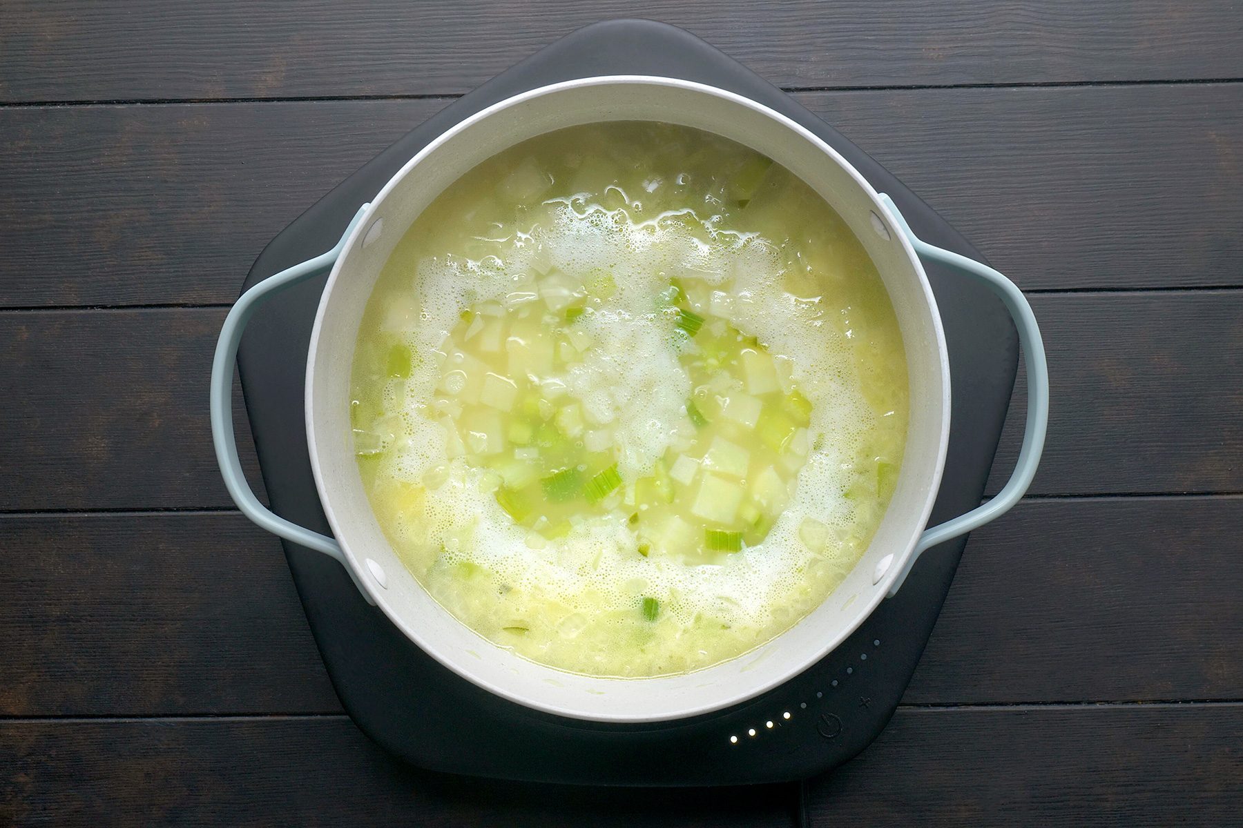 A pot of bubbling soup on a dark wooden surface, featuring diced potatoes and green vegetables in a creamy broth.