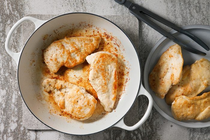 overhead shot of two plates one with cooked chicken breast fillets in a white skillet and another plate with more cooked chicken breast fillets, The skillet is on a light grey background with a pair of black tongs to the top right, The chicken fillets are slightly browned;