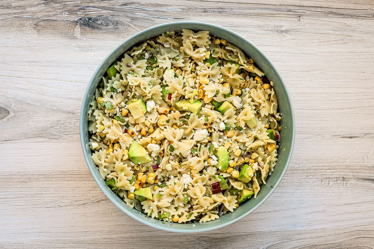 Preparing Taste of Home's Mexican street corn pasta salad with bow tie pasta, avocado cubes, cotija cheese, grilled corn kernels, and red onions.