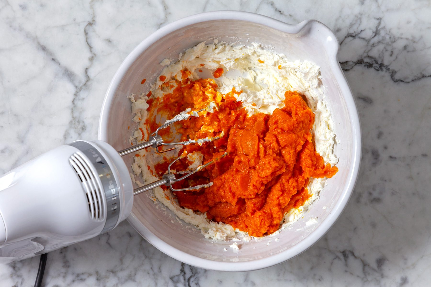Overhead shot of a white bowl containing a mixture of cream cheese and pumpkin puree, An electric mixer is partially submerged in the mixture.