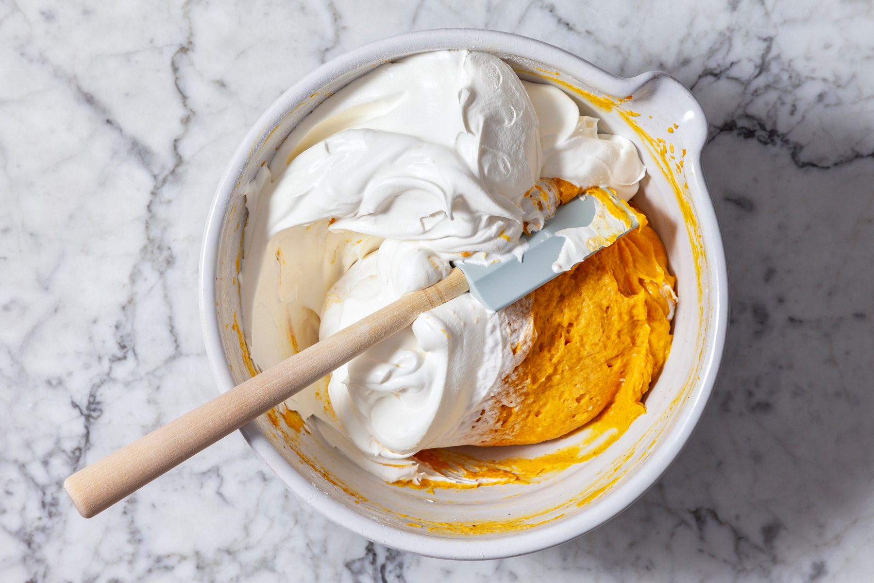 Overhead shot of a white bowl containing two distinct layers of mixture, The bottom layer is a vibrant orange color, likely a pumpkin-based filling, The top layer is white and fluffy, suggesting it is whipped cream, A spatula is gently folding the two layers together, creating a marbled effect.