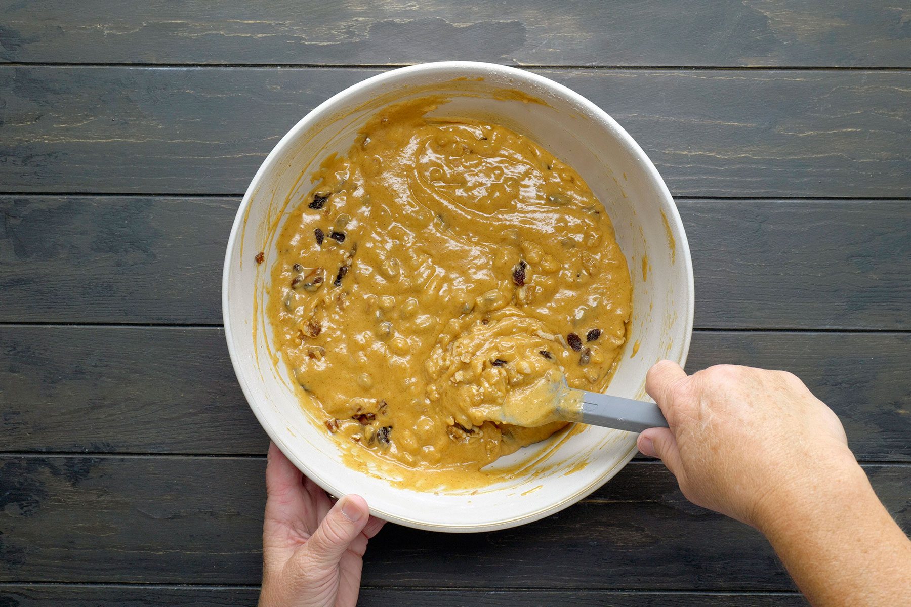 A person mixes cookie dough with chocolate chips in a large white bowl using a spatula. The hands hold the bowl on a dark wooden table.