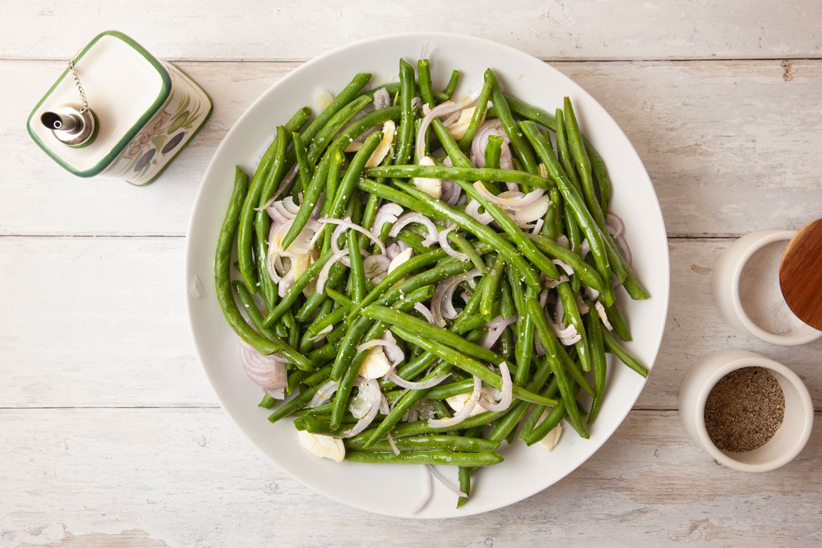 Step 1 of Taste of Home Roasted Green Beans is to combine the green beans, shallots and garlic. Drizzle the vegetables with olive oil, sprinkle them with the salt and pepper, and toss them until they’re well coated. Divide the beans between two 15x10-inch baking pans, coating with cooking spray.