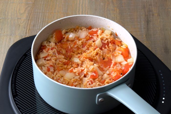 The saucepan contains a mixture of rice, chopped tomatoes, and chopped onions, The rice is a white color and the tomatoes and onions are a vibrant red color, The mixture is slightly oily
