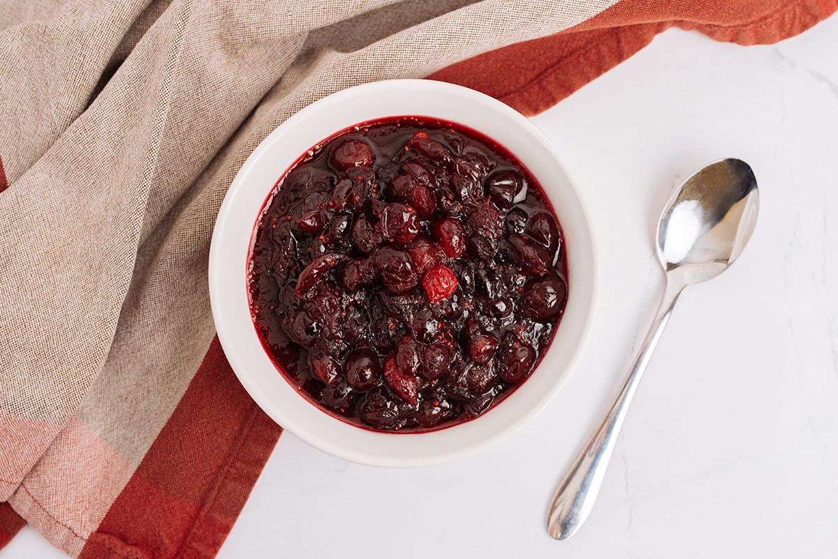 Cranberry sauce in a white bowl with a pink and maroon cloth napkin in the background with a silver spoon next to it