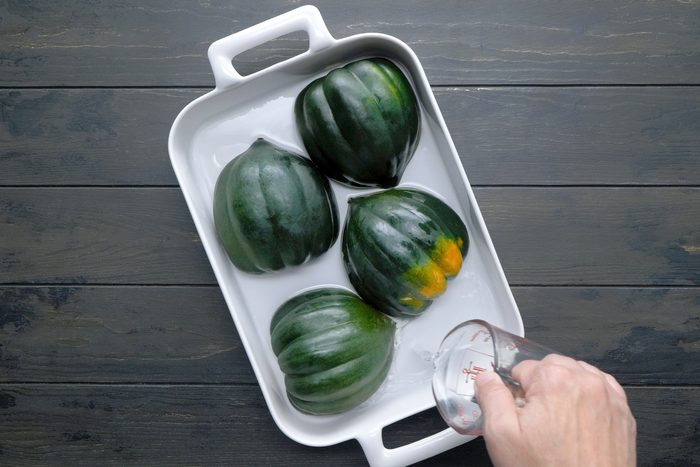 overhead shot of a white baking dish filled with acorn squash, The acorn squash are green with orange patches and are submerged in water, A person's hand is pouring water from a measuring cup into the dish;