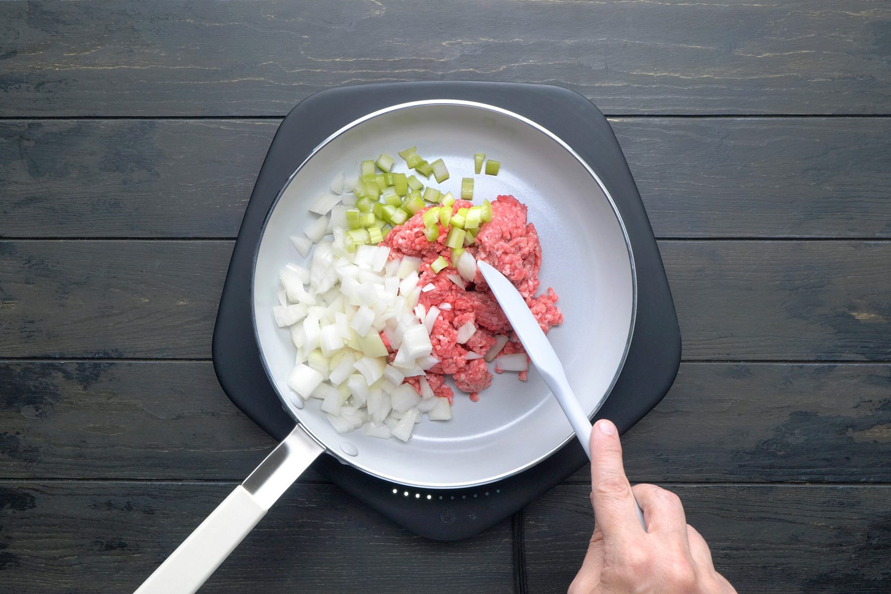 overhead shot of A white skillet is filled with a mixture of ground meat (likely beef or turkey), chopped onions, and chopped celery, The meat is raw and the vegetables are bright green and white, A spatula is being used to stir the ingredients together;
