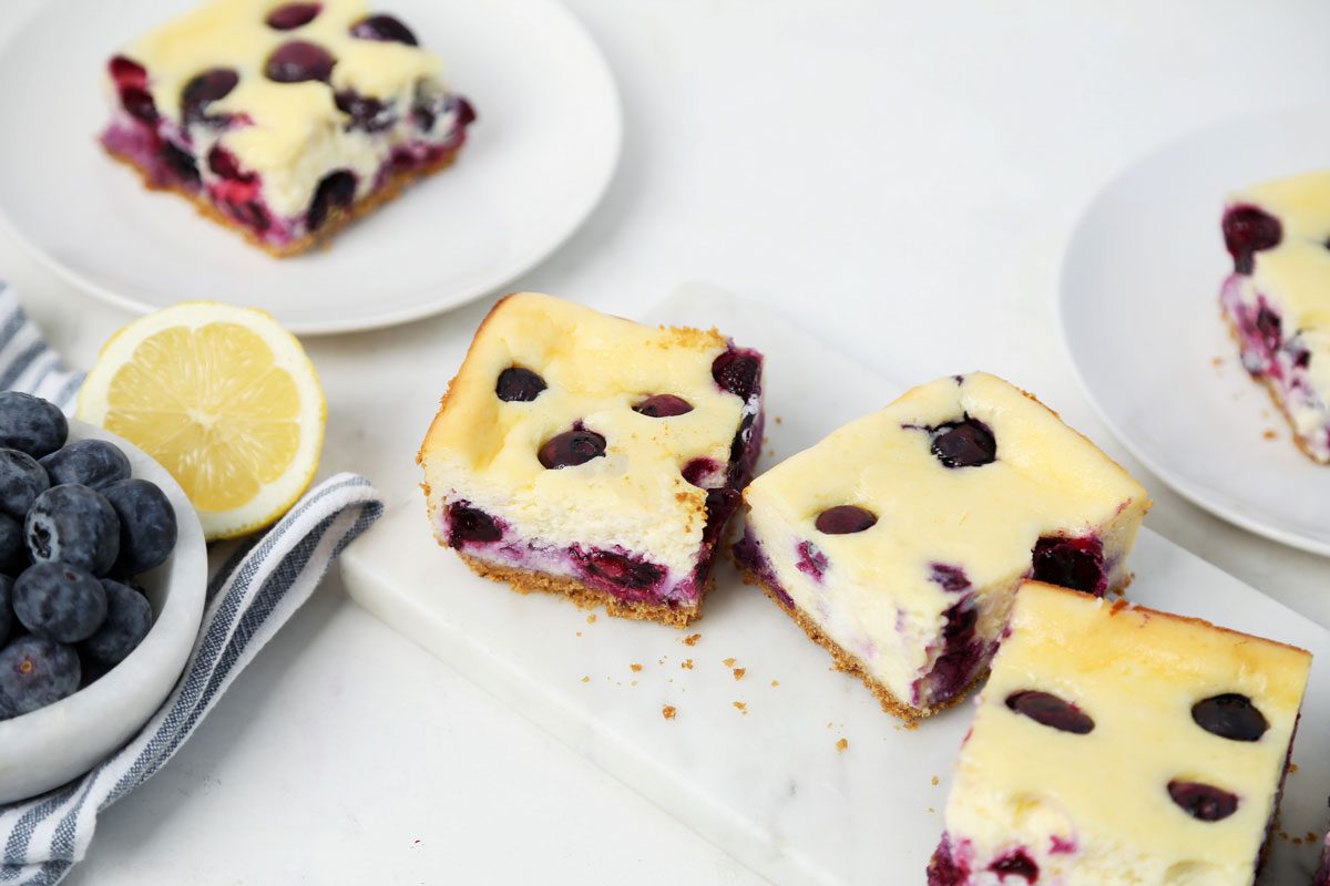 Close up of Taste of Home's Blueberry Cheesecake Bars sliced and served on a platter on a white wooden surface.