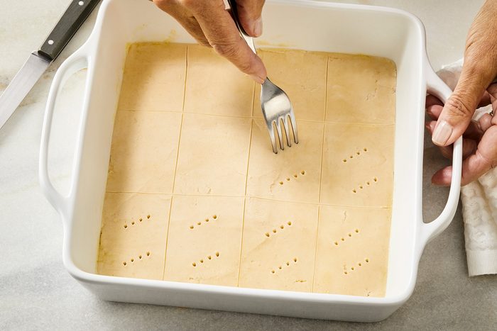 3/4th shot of Using a knife to score batter into 2 in squares; Using a fork to prick a pattern onto the top of each rectangle in a white baking dish;