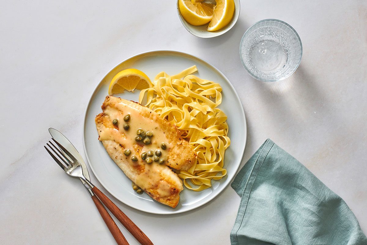 Tilapia fillet served with pasta and lemon butter sauce on a serving plate with a lemon wedge on the side