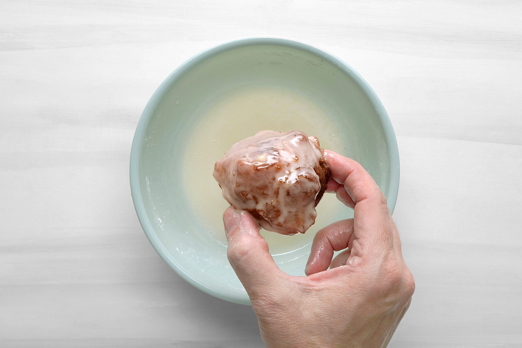 overhead shot of a hand holding a glazed donut that is being dipped into a bowl of glaze; the bowl is light blue and sits on a white surface; the hand is holding the donut by the bottom with the fingers curled around it;