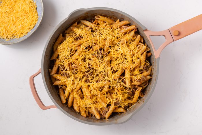 Bacon cheeseburger pasta being finalized with shredded cheddar cheese on top.
