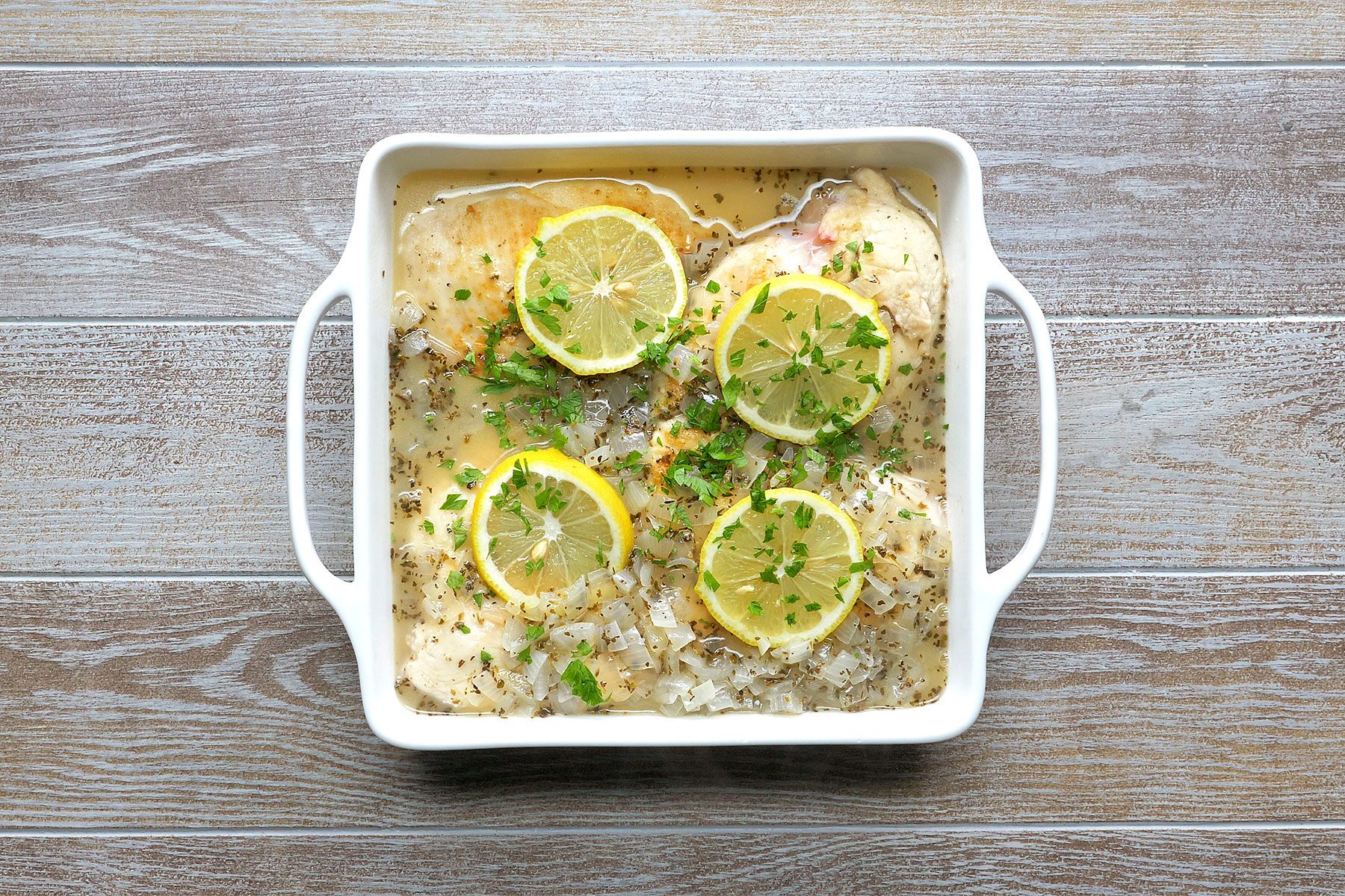 overhead shot of a white baking dish filled with chicken breasts, lemon slices, chopped onions, and herbs, all submerged in a clear broth, resting on a wooden surface