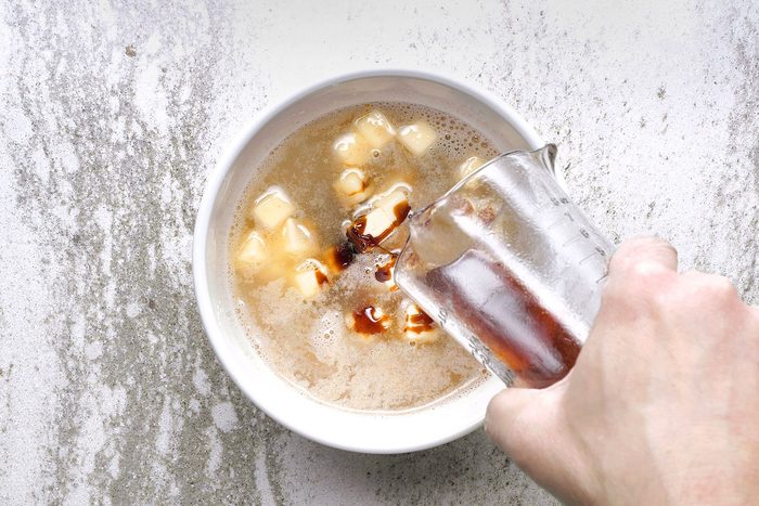 A hand pouring liquid from a glass measuring cup into a bowl containing a foamy mixture with small yellow cubed pieces, likely butter. The background is a textured light surface.