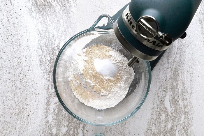 A stand mixer with a glass bowl containing flour and sugar on a textured white and gray surface.