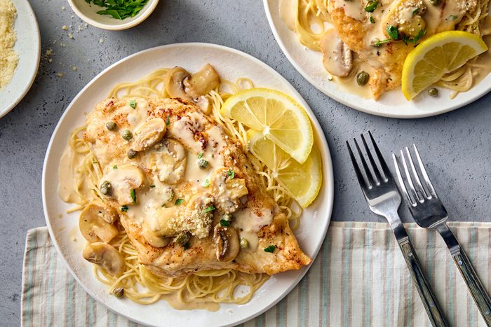 Top view shot of Cheesecake Factorys Chicken Piccata; serve on plates; forks; napkin; cheese; lemon slices; grey surface.