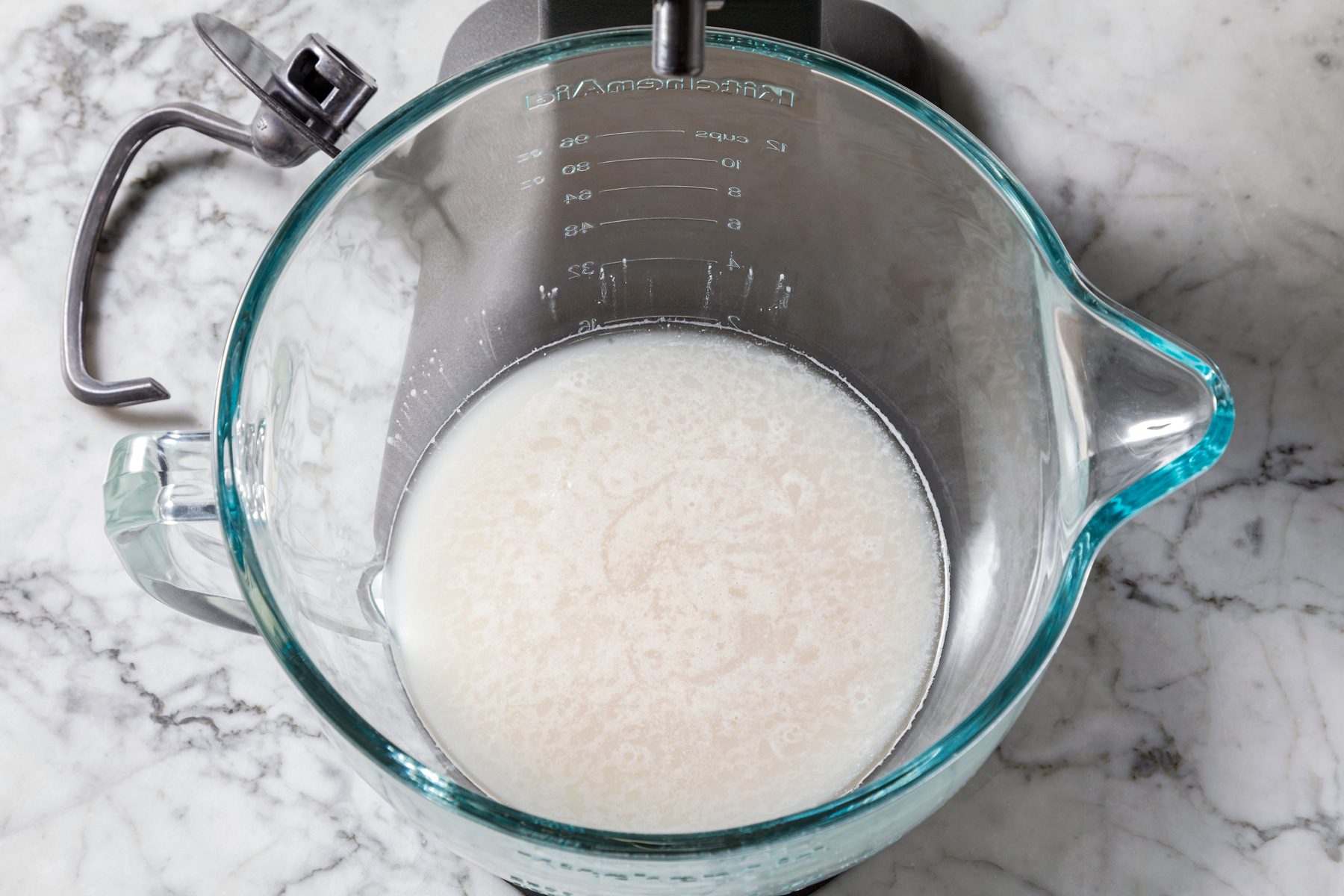 Overhead shot of a stand mixer bowl with hook attachment, warm water and yeast proofing on a marble surface for 5 minutes.
