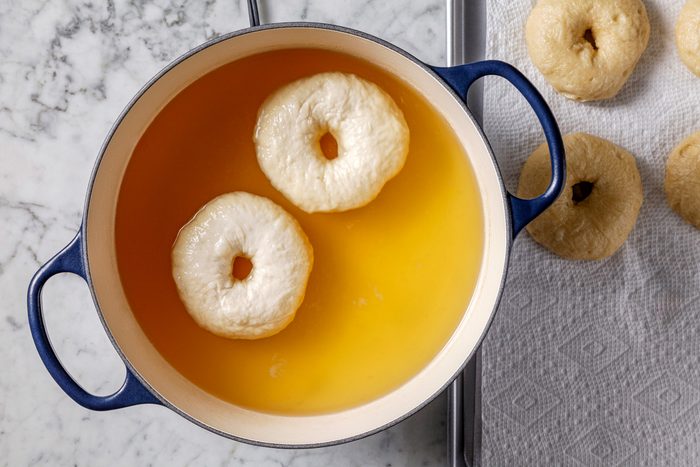 Overhead shot of a Dutch oven filled with water and honey, brought to a boil; adding bagels two at a time, cooking for 45 seconds per side, then removing with a slotted spoon to drain on paper towels, on a marble surface.
