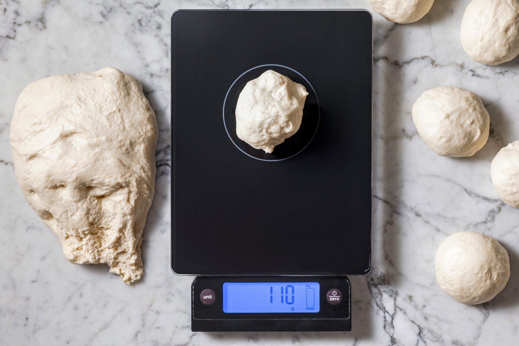 Overhead shot of dough on a weighing scale, with dough shaped into smooth balls on a marble surface.