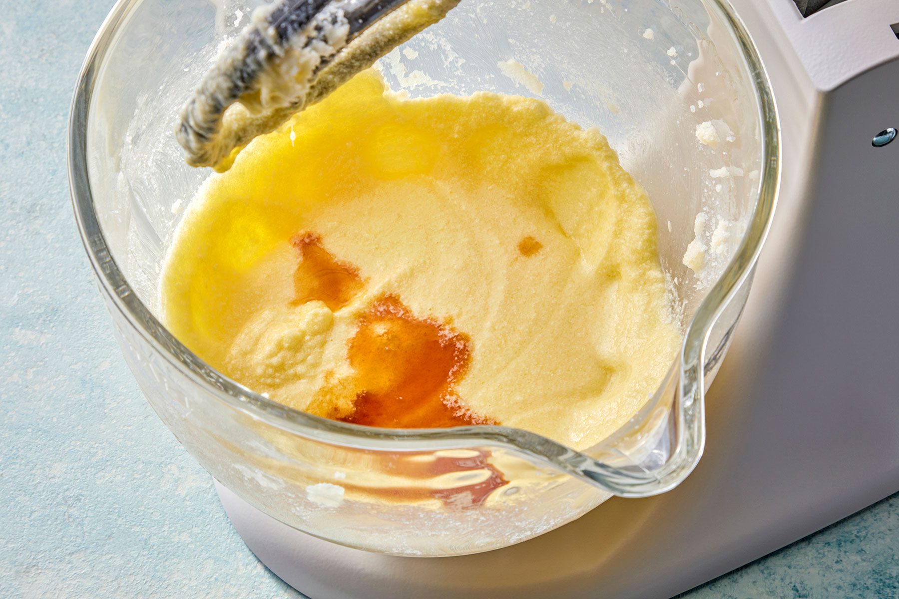 A close-up of a stand mixer bowl with creamed butter and sugar, partially mixed. The mixture appears light and fluffy with a splash of vanilla extract added. The background is a light blue countertop.