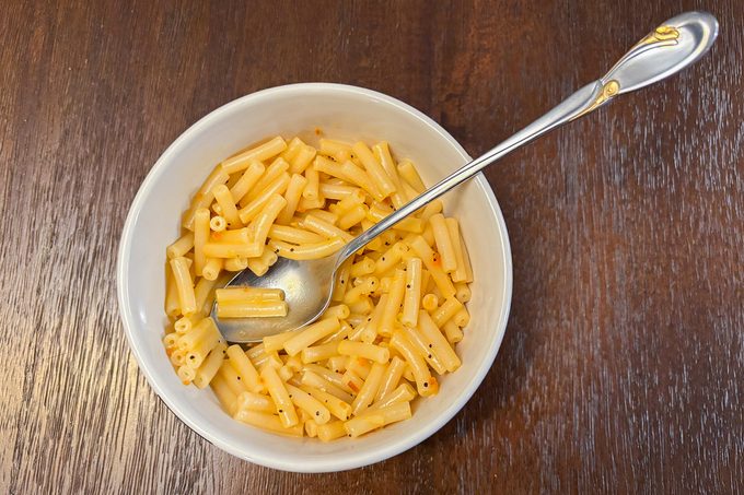 A white bowl filled with cooked macaroni pasta on a wooden table. A silver spoon is resting in the bowl. The macaroni is lightly coated with a yellowish sauce.