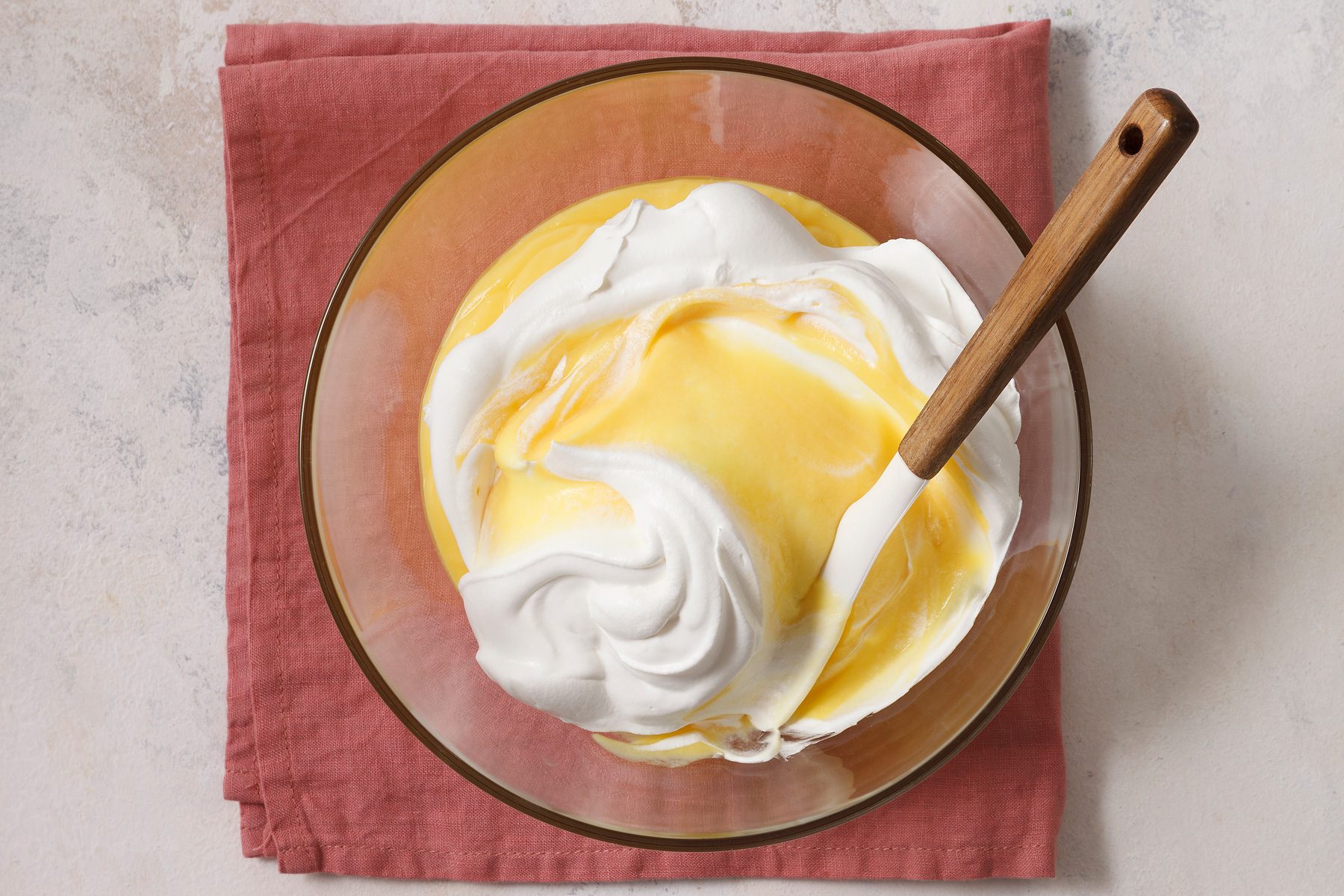 Overhead shot of same bowl; gently fold one carton whipped topping into pudding; spatula; napkin; marble surface.