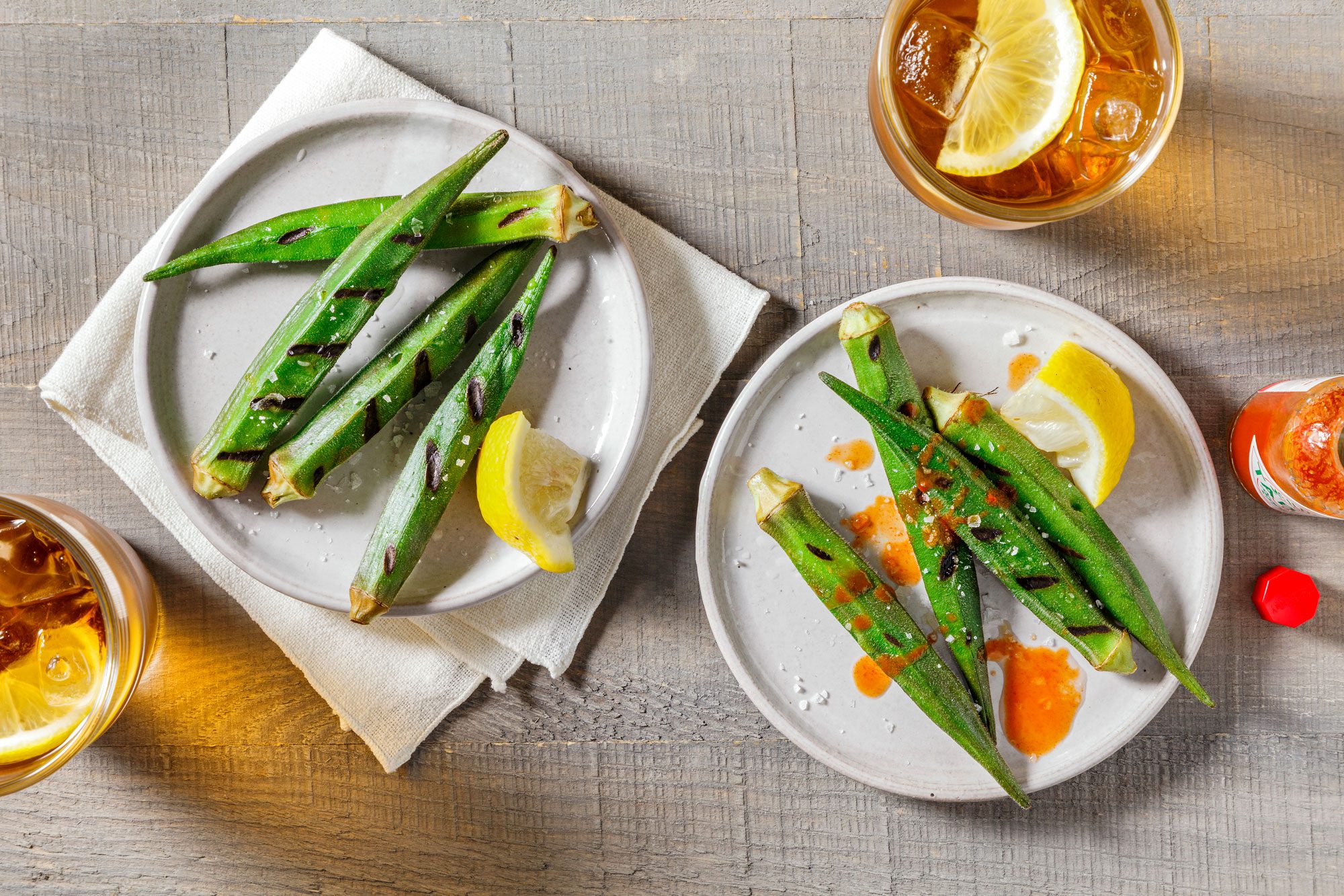 Overhead shot of Grilled Okra; serve hot on plates; garnish with lemon juice and salt; drinks; napkin; wooden surface;