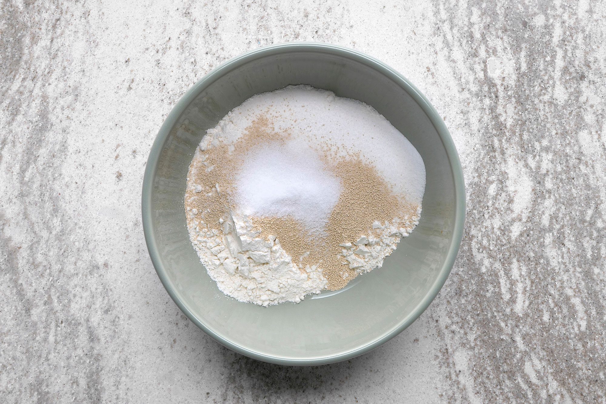 overhead shot of a light grey bowl with a white rim sits on a grey and white speckled surface; inside the bowl is a mixture of flour, sugar, and yeast