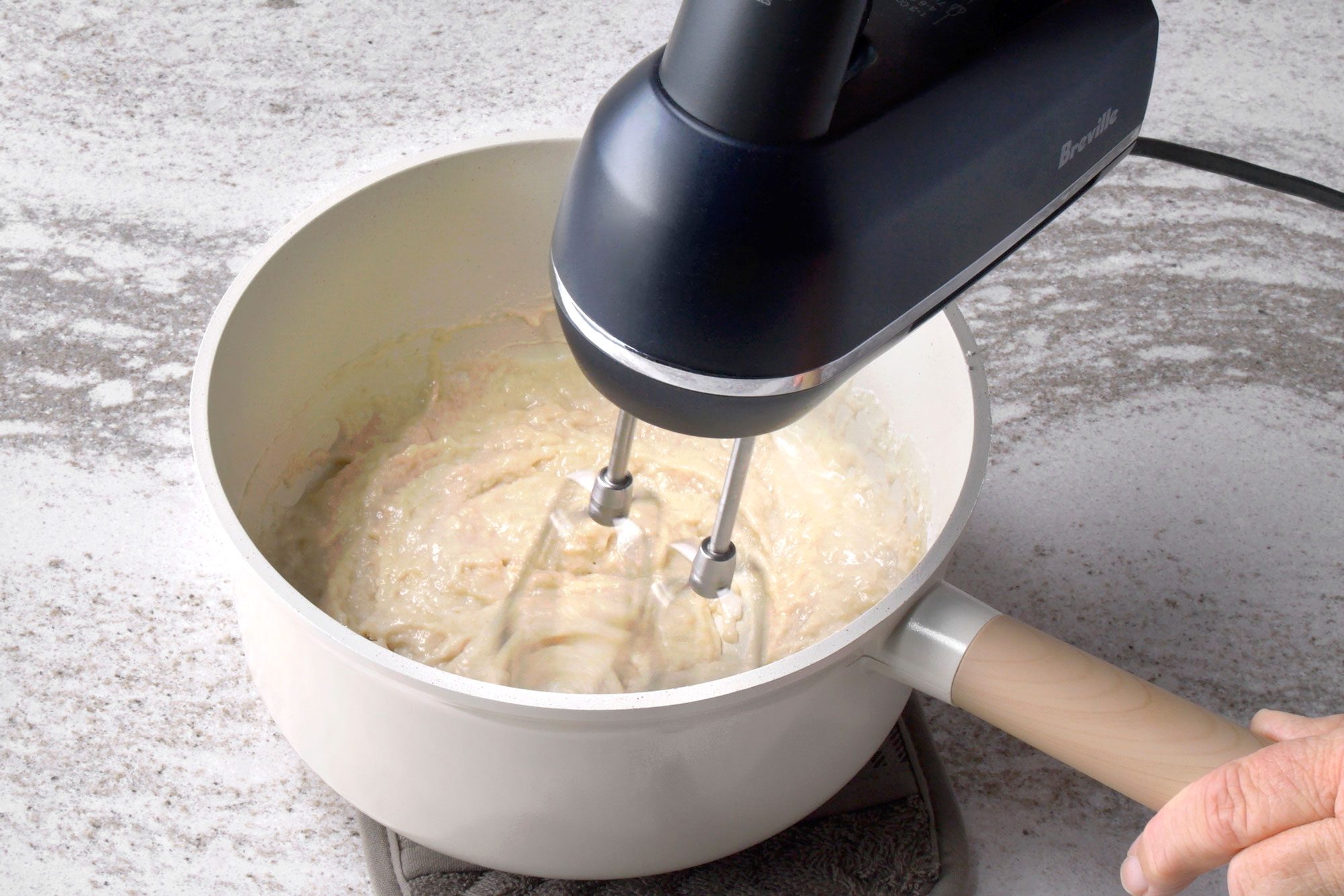 3/4th shot of a hand is holding a handle of a hand mixer, which is mixing a batter in a white bowl; the batter is thick and creamy and the bowl is sitting on a grey and white countertop; the hand mixer is black, the mixer is plugged in and the cord is laying on the countertop