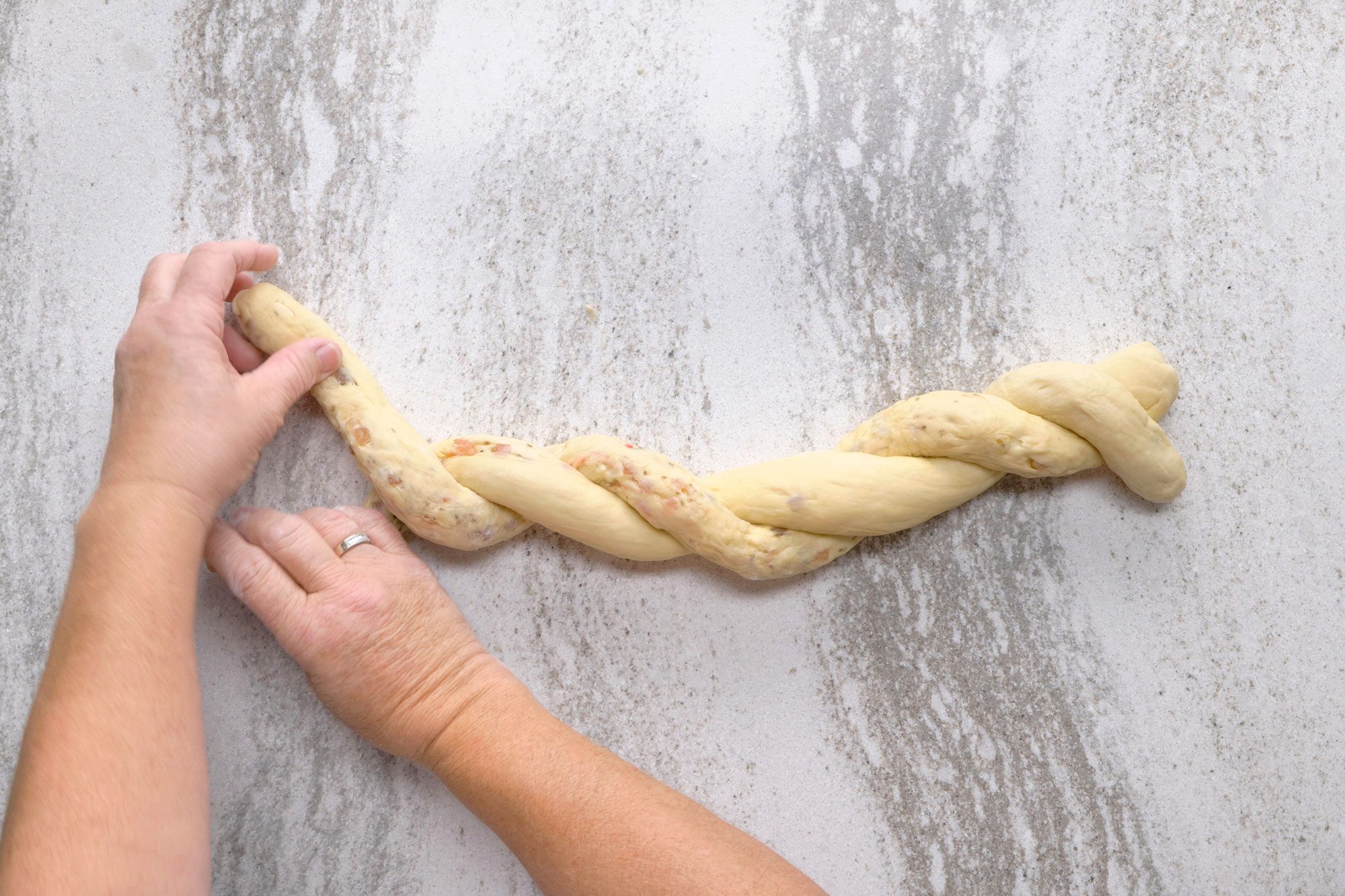 overhead shot of a person is braiding a loaf of dough; they are using both hands to shape the dough and the dough is on a white surface, possibly a countertop; the dough is braided in a three-strand pattern and looks like it will be braided bread, there are visible specks of something in the dough, perhaps raisins or nuts;