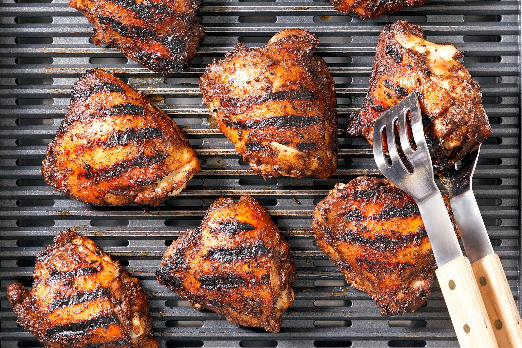 overhead shot of a close-up view of a grill with several pieces of grilled chicken, the chicken pieces are arranged in a circle around the center of the grill; a set of grilling tongs is positioned in the center of the image; the tongs are made of metal with wooden handles, they are holding a piece of chicken; the chicken is cooked to a golden brown and has visible grill marks; the grill surface is dark gray and has a pattern of raised lines
