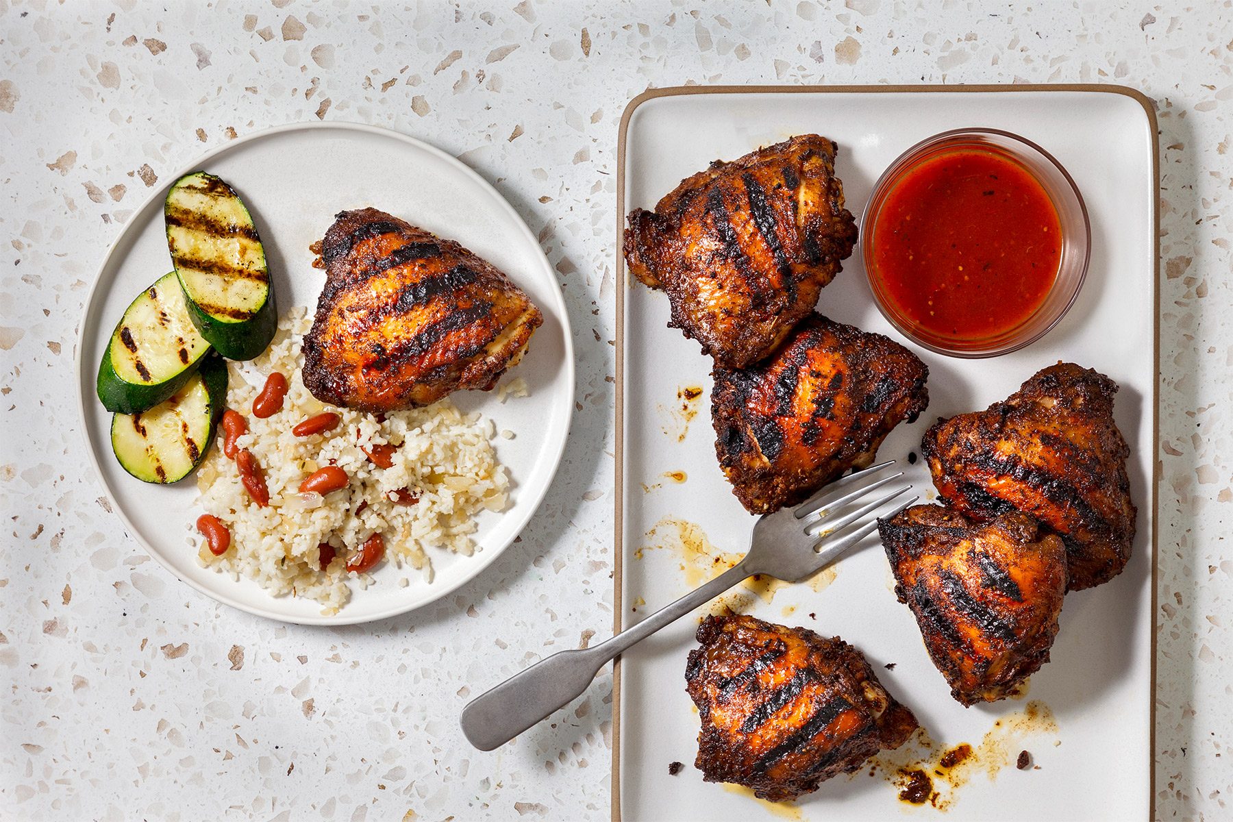 overhead shot of plate of food with grilled chicken, rice, red beans and zucchini, and a plate of grilled chicken with a small bowl of red sauce, a fork, and a few drops of sauce on the plate, all laid out on a white countertop with brown flecks