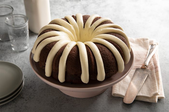 A chocolate bundt cake decorated with white icing drizzled in a radial pattern. It's placed on a cake stand beside a stack of plates, a knife, and napkins. Two glasses and a bottle are in the background on a gray surface.