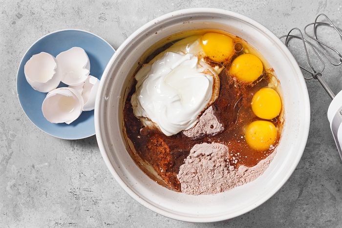 overhead shot of a white mixing bowl sits on a grey countertop; the bowl is filled with the ingredients for a cake batter or other baked good; the ingredients include a dollop of sour cream, four egg yolks, a brown liquid, and some cocoa powder; a blue plate sits to the left of the bowl; it holds three broken eggshells; a silver hand mixer sits on the right side of the bowl