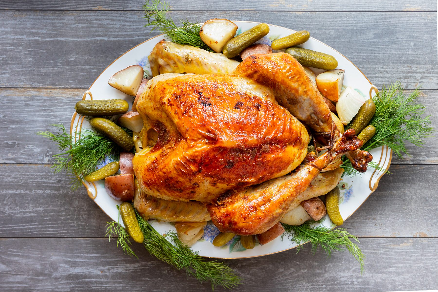 A roasted turkey on a platter, garnished with dill, small roasted potatoes, and pickles, displayed on a wooden table.
